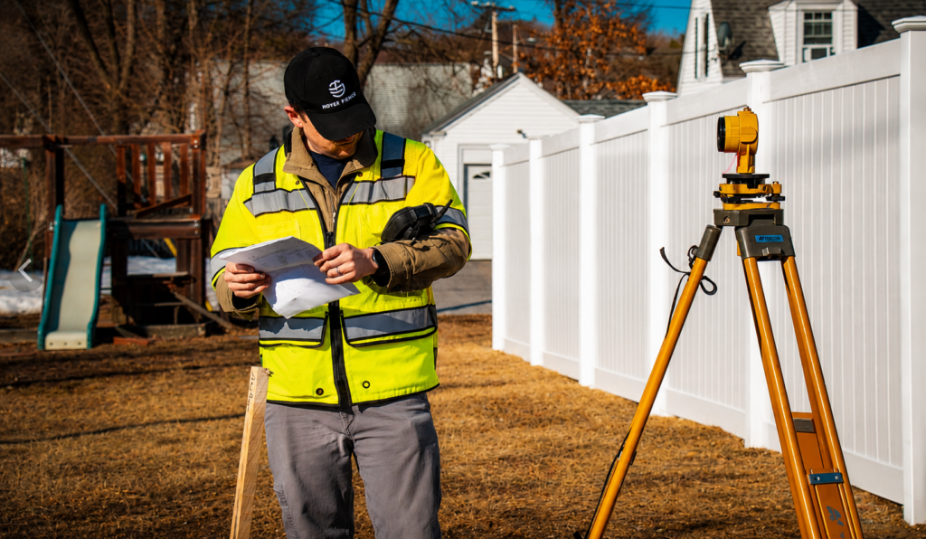 Professional land surveyor reviewing site plans beside a tripod-mounted total station during a residential construction project.