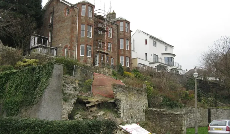 Retaining wall failure example, a collapsed stone retaining wall below houses on a sloping site